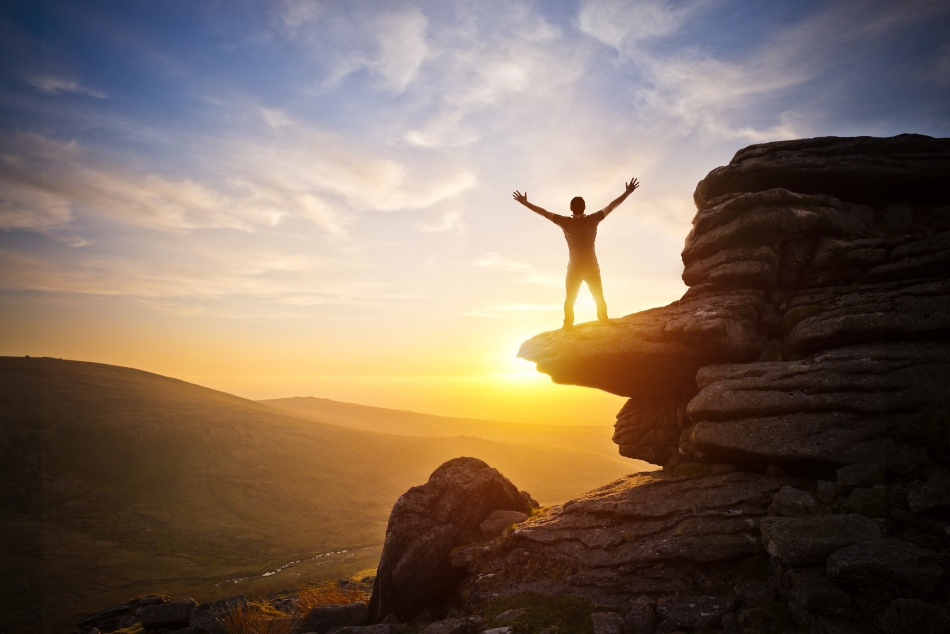 A person standing on a clifftop at sunrise with both arms raised